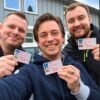 Trois hommes souriants montrant leur permis de conduire devant un bâtiment après leur réussite à l’examen.