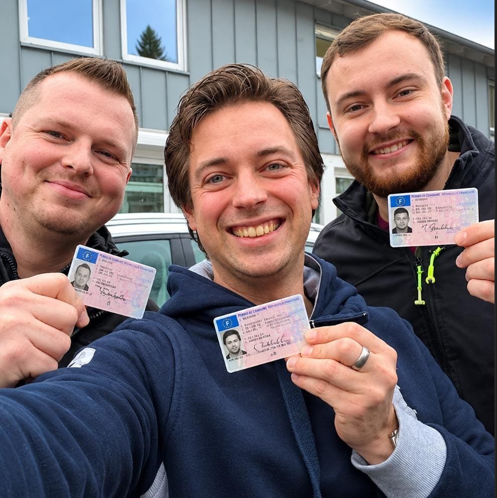 Trois hommes souriants montrant leur permis de conduire devant un bâtiment après leur réussite à l’examen.
