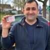 Homme souriant montrant son permis de conduire devant une voiture après sa réussite à l’examen.