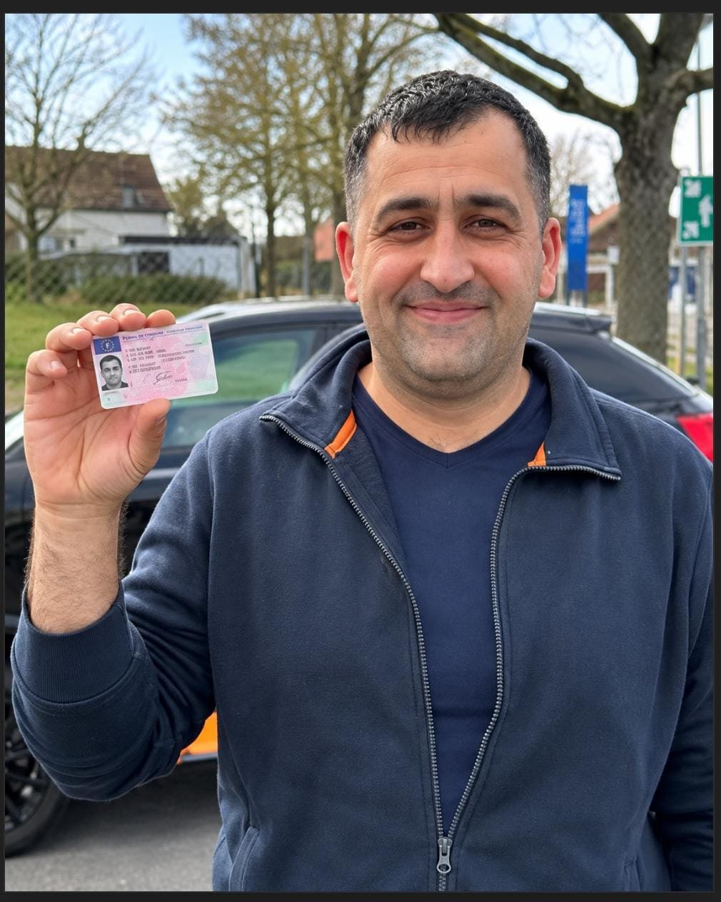 Homme souriant montrant son permis de conduire devant une voiture après sa réussite à l’examen.